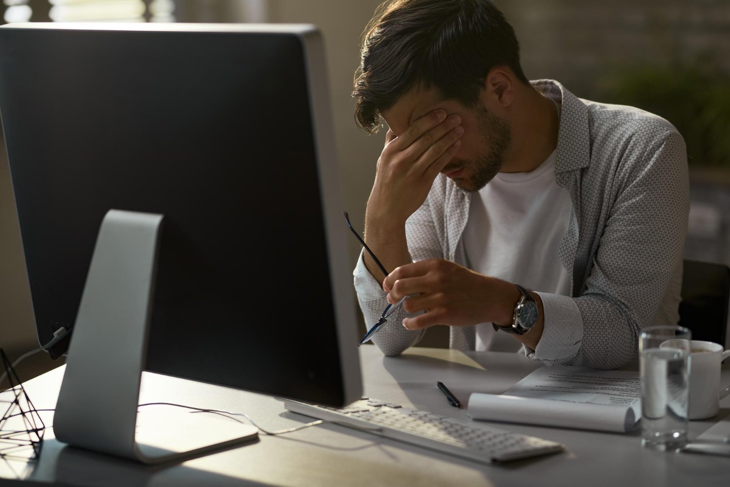 Homem sentado à mesa de trabalho, com óculos na mão, esfregando os olhos em sinal de exaustão, ilustrando esgotamento por burnout